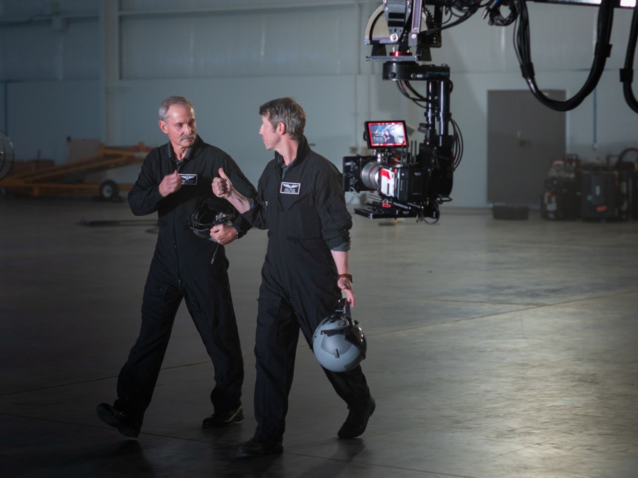 Two men walking through a jet hangar, carrying helmets. There is a movie camera following them on a jib.
